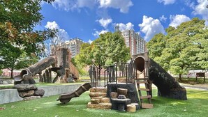 Un parque infantil con toboganes y estructuras para escalar, rodeado de árboles y un edificio al fondo.