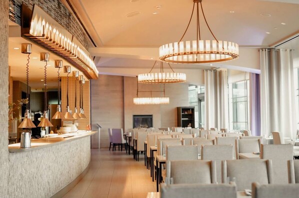 Interior modern d'un restaurant amb una barra llarga, diverses làmpades penjants i files de taules i cadires.