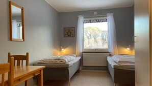 Ett litet, väl upplyst rum med två enkelsängar, en trästol, ett bord och ett fönster med gardiner.