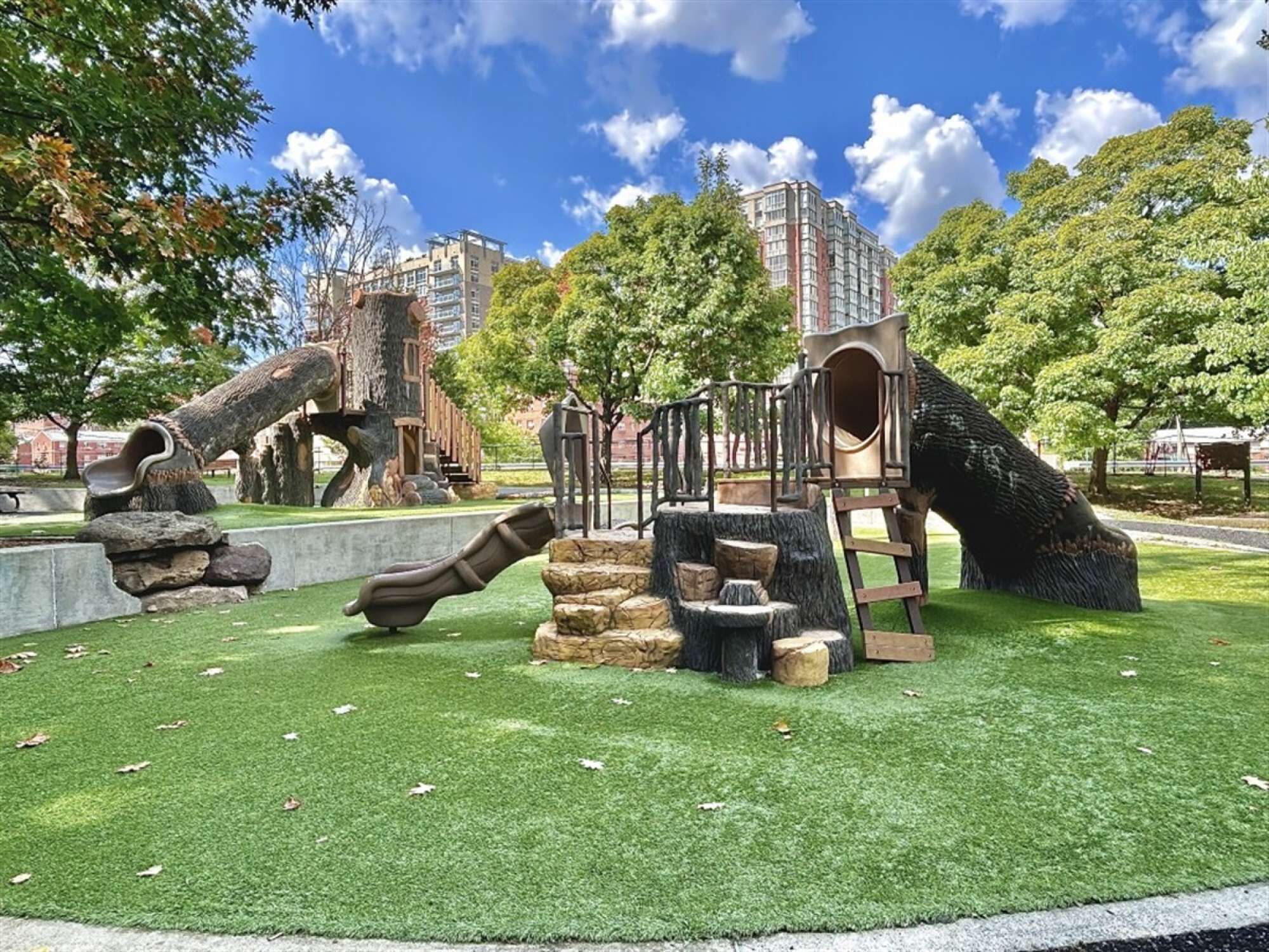 Un parque infantil con toboganes y estructuras para escalar, rodeado de árboles y un edificio al fondo.