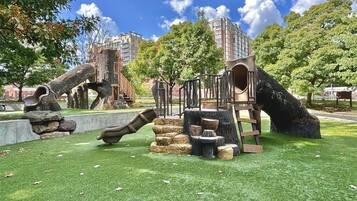 Un parque infantil con toboganes y estructuras para escalar, rodeado de árboles y un edificio al fondo.