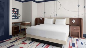 A hotel room with a bed, bedside table, chair, desk, and a wall clock.