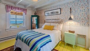 Une chambre à coucher avec un lit, une fenêtre donnant sur un beau paysage, une commode et un mur orné d’un motif floral.