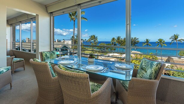 A balcony with a glass table set for dining, wicker chairs, and a view of the ocean and palm trees. - #510-Ocean and Sunset Views! Steps from It All! (Kihei)