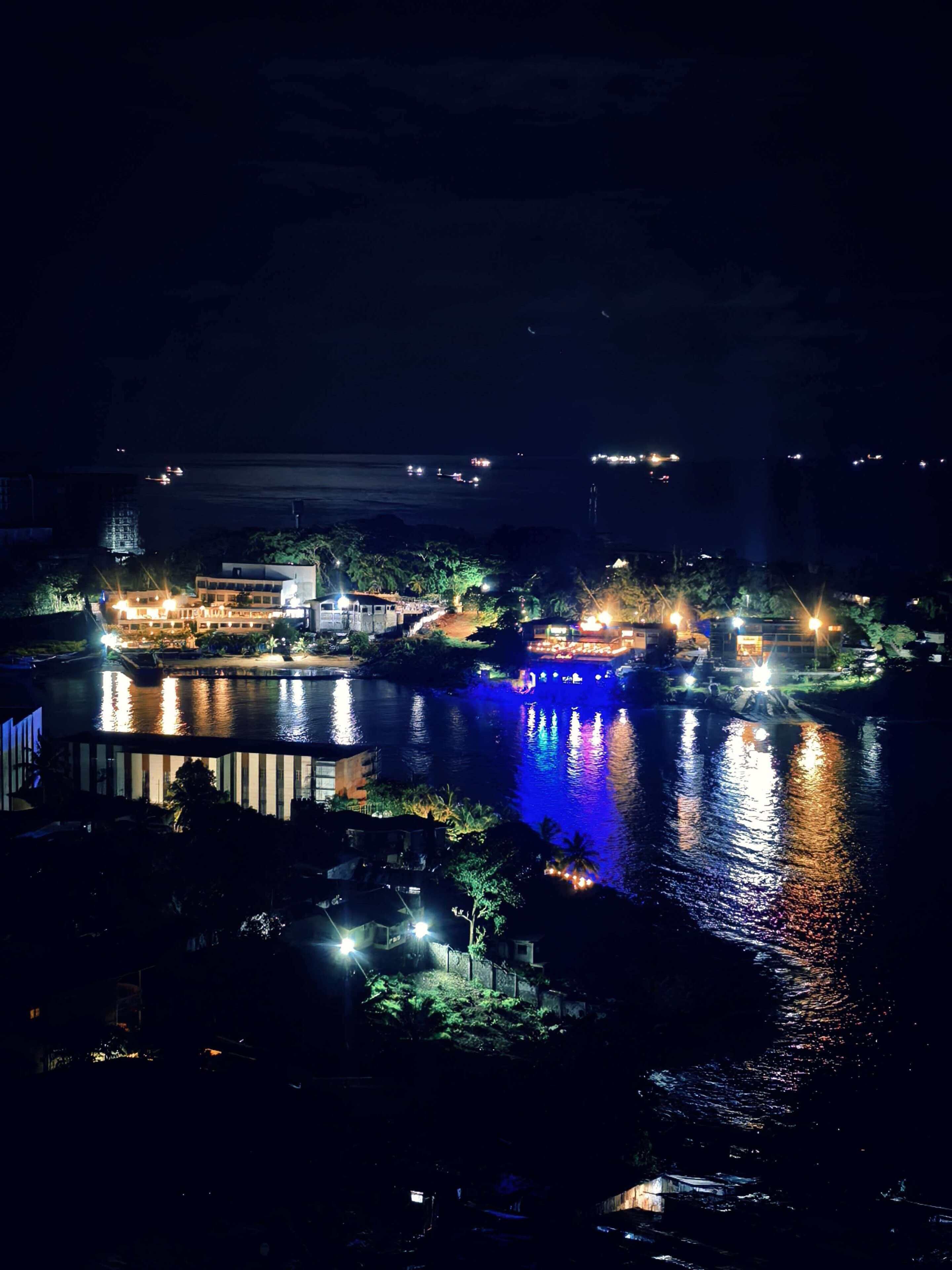 Vista noturna de uma área à beira-mar, com edifícios iluminados, um corpo d'água refletindo a luz e uma paisagem urbana ao longe.
