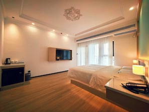 A hotel room with a bed, bedside table, lamp, TV, and a desk with a kettle and cups. - bintumani hotel (Freetown)