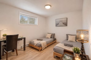 Quarto pequeno e bem iluminado, com uma escrivaninha, duas camas de solteiro, uma cadeira, uma mesa com uma planta e um abajur.