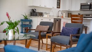 A modern living room with a glass coffee table, two grey armchairs, a blue sofa, and a kitchen in the background with white cabinets and appliances. - Fully Furnished Sandy Home | Monthly Stays (Sandy)