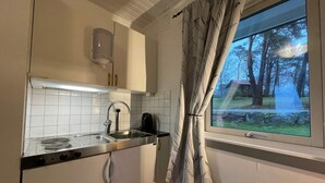 Una cocina compacta con armarios blancos, un lavamanos de acero inoxidable y una ventana con vistas a árboles y una casa.