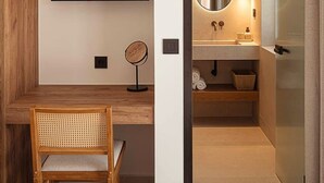 Una habitación de hotel moderna con un escritorio de madera, un televisor de pantalla plana, un espejo redondo y un baño con lavabo y espejo.