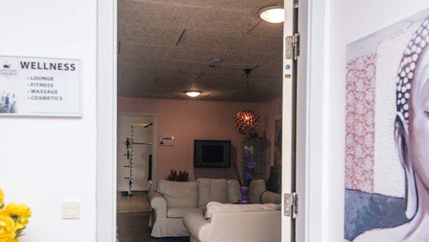 A room with a white couch, a small table, and a wall-mounted shelf. There is a sign on the wall listing amenities such as a lounge, fitness area, massage, and cosmetics. - 4 Person Holiday Park Home in Romo (Rømø)