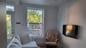 Ein Zimmer mit einem weißen Sofa, einem Sessel, einem an der Wand befestigten Fernseher und einem Fenster mit Blick auf Gebäude und Bäume.