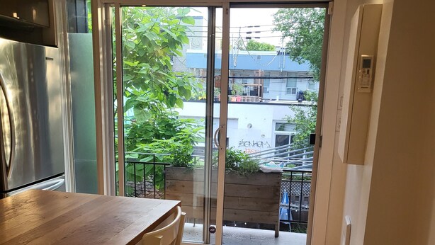 Eine Küche mit einem Holztisch und Stühlen, einem Kühlschrank und einer Glastür, die auf einen Balkon mit Blick auf Gebäude und Grünflächen führt.