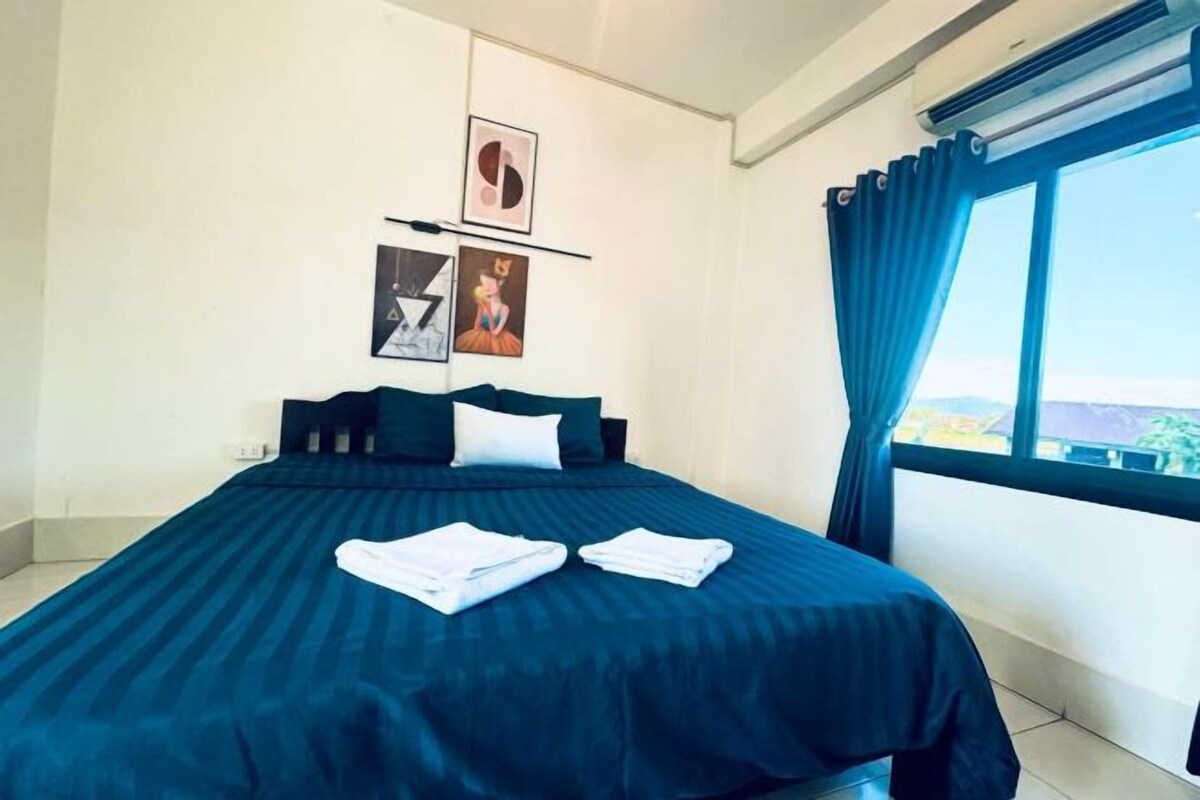 Un lit bien fait, recouvert d’une couvre-lit bleu foncé, et deux serviettes blanches pliées sont posées dessus. Deux tableaux encadrés sont accrochés au mur, au-dessus du lit.