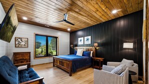 A bedroom with a wooden ceiling, a bed with blue bedding, a wooden dresser, a striped armchair, and a window with blinds. - Grand Forge Lodge - 16 Bedrooms in the MIDDLE of Pigeon Forge! Loaded! (Pigeon Forge)