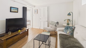 Una sala de estar con una televisión de pantalla plana sobre un mueble de madera, una mesita de centro, un sofá, una silla y una mesita auxiliar con una lámpara.