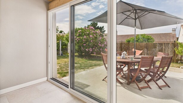 Un patio con una mesa y sillas, una sombrilla y un ventanal corredera que da a un jardín.