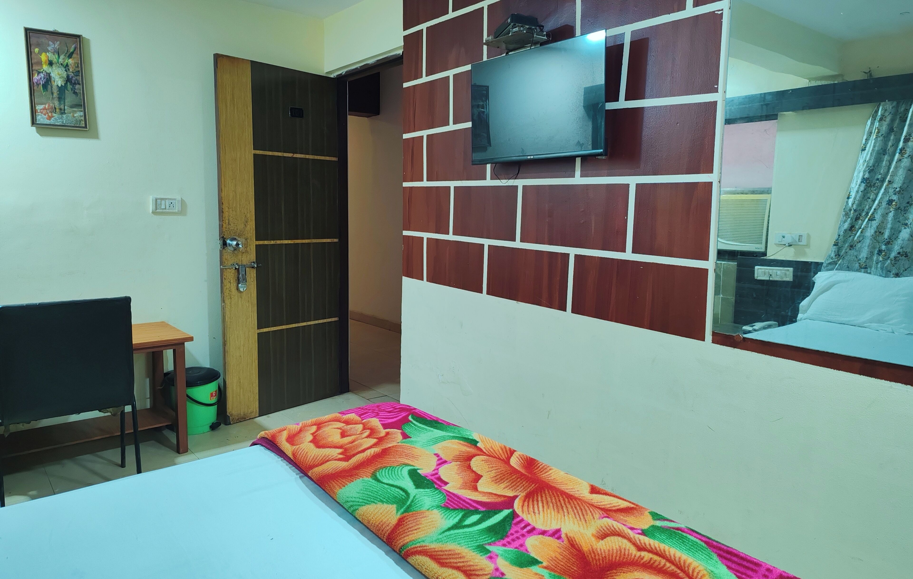 A room with a bed, a TV mounted on a brick-patterned wall, a wooden door, and a small table with a bucket.