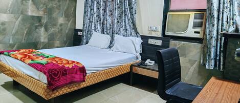A hotel room with a bed, a bedside table with a phone, a chair, and an air conditioning unit.