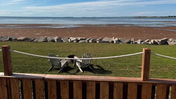 A wooden deck with a rope barrier, a grassy area, and a view of a beach with a rocky shoreline and calm water. - Schurman’s Shore Cottage #3 (Travellers Rest)