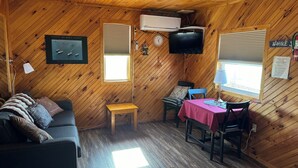 A cozy cabin with a wooden interior, a sofa, a small table, a TV, and a window with blinds. - Schurman’s Shore Cottage #3 (Travellers Rest)