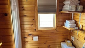 A compact wooden cabin bathroom with a toilet, sink, and a window with blinds. - Schurman’s Shore Cottage #3 (Travellers Rest)