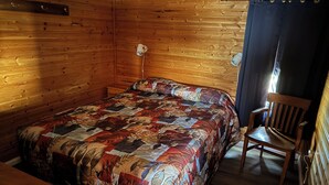 A wooden cabin room with a bed, a chair, a nightstand, and a lamp. - Schurman’s Shore Cottage #3 (Travellers Rest)