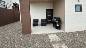 Ein überdachter Außenbereich mit Grill, zwei schwarzen Sitzgelegenheiten mit Kissen und einer schwarzen Tür.