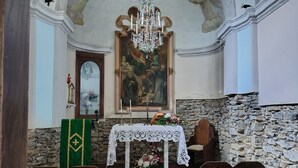 A church interior with a stone wall, wooden pews, a white altar table, and a large religious painting. - The Bishop's House (Licciana Nardi)