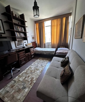 A room with a sofa, a desk, a computer, a bookshelf, and a window with curtains.