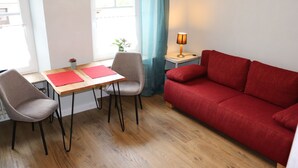A room with a wooden floor, a red sofa, a wooden table with two chairs, a lamp, and a window with curtains.