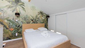 A bedroom with a bed, white bedding, a mural of tropical plants, and two wall-mounted lamps. - Dify Jungle - Villeurbanne (Villeurbanne)