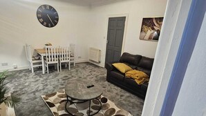 Een woonkamer met een ronde salontafel, witte stoelen, een zwarte bank, een klok en een radiator.