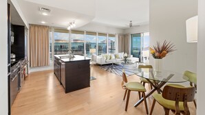 A modern living room with a glass dining table, a sofa, and a kitchen area with a large island. - LUXURY 2-Bed Condo, Kitchen, Wi-Fi, Parking, W/D, AC, LEGAL Monthly Rental! (Honolulu)
