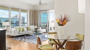 A modern living room with a sofa, coffee table, and chairs, large windows with a view of buildings, and a ceiling fan. - LUXURY 2-Bed Condo, Kitchen, Wi-Fi, Parking, W/D, AC, LEGAL Monthly Rental! (Honolulu)