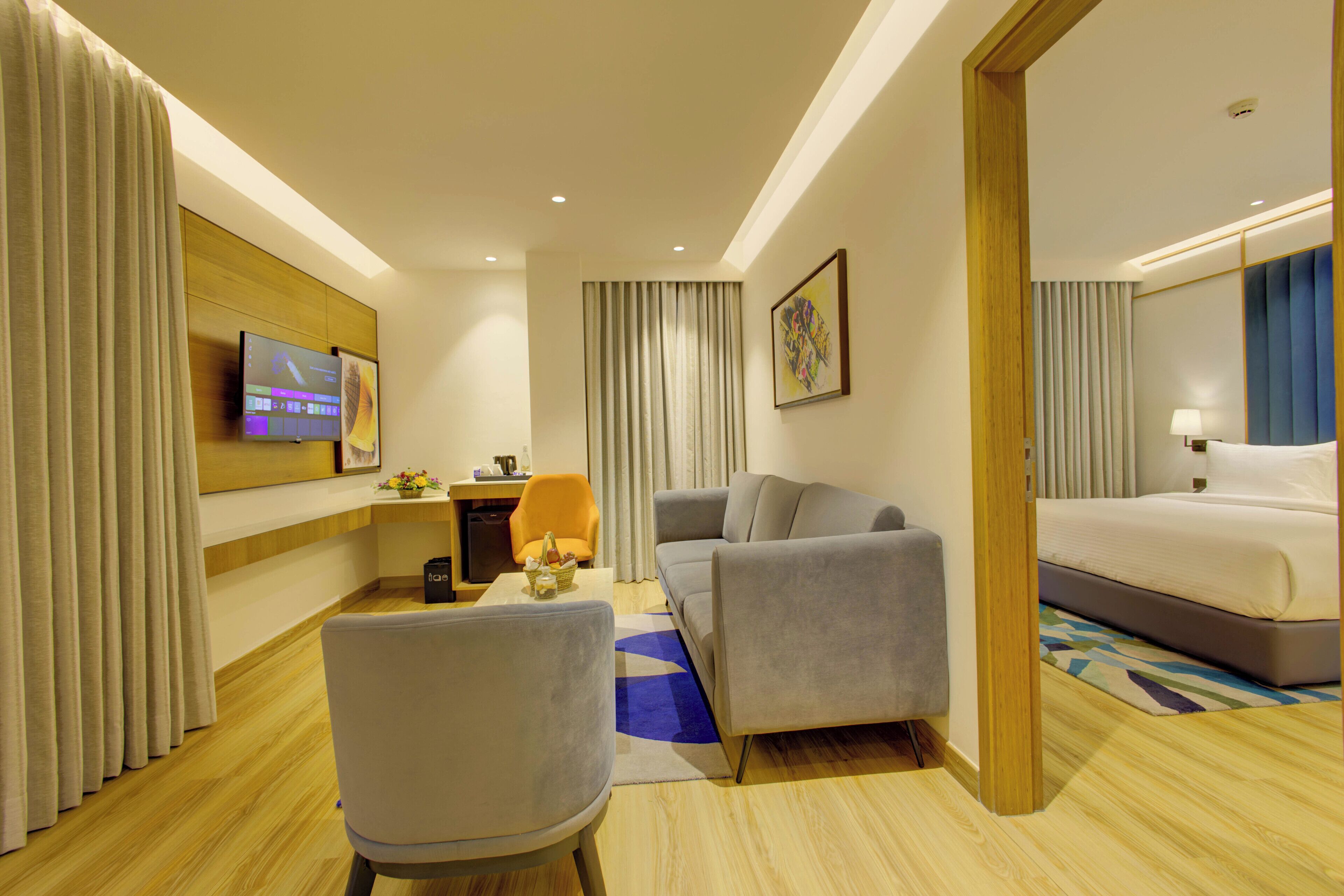A hotel room with a bed, a sofa, a chair, a desk with a computer, and a TV on the wall.
