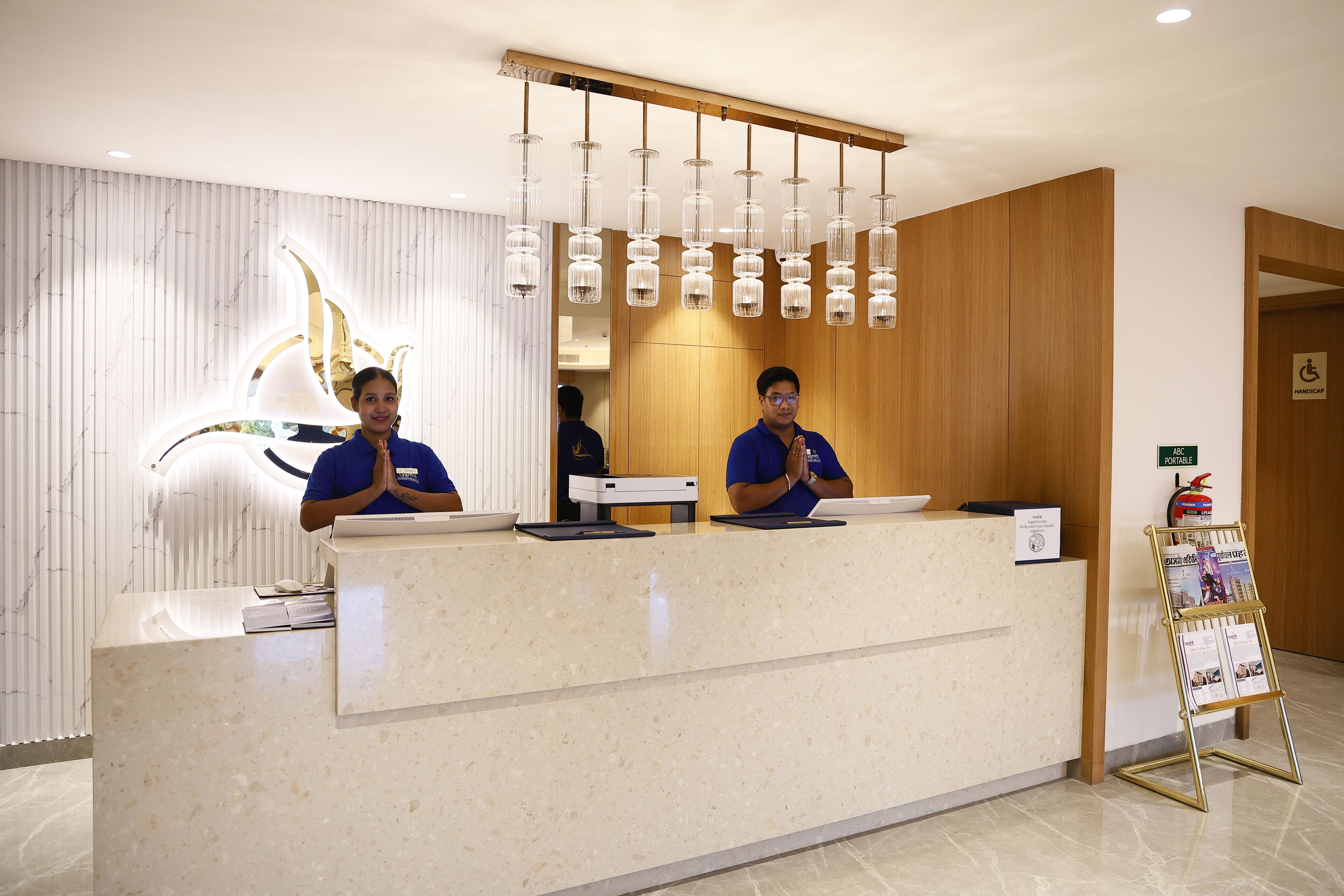 Un comptoir d’accueil d’hôtel avec deux employés, un mur en marbre orné d’un motif de lotus et un panneau indiquant une zone accessible aux personnes handicapées.