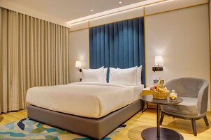 Una habitación de hotel con una cama grande, una silla, una mesita con una cesta y un tarro de cristal, y una pared con un panel azul y cortinas.