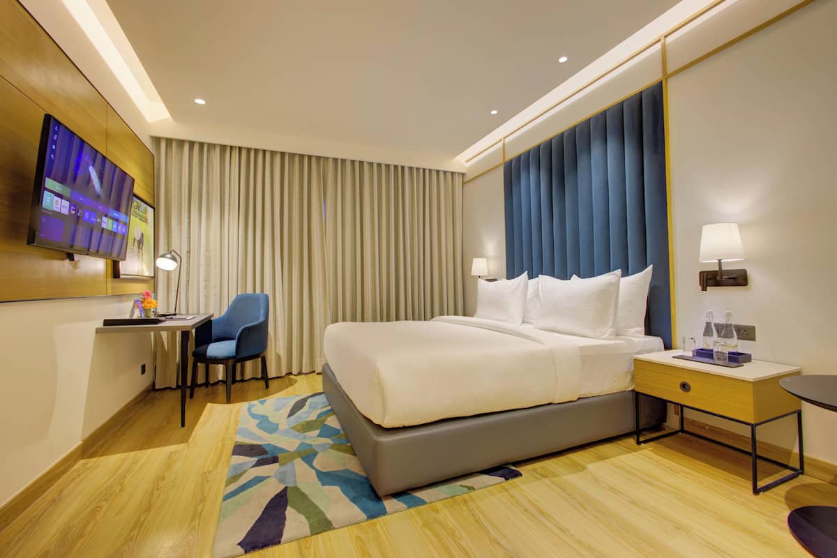 A hotel room with a large bed, a desk with a chair, a flat-screen TV, and a large window with curtains.