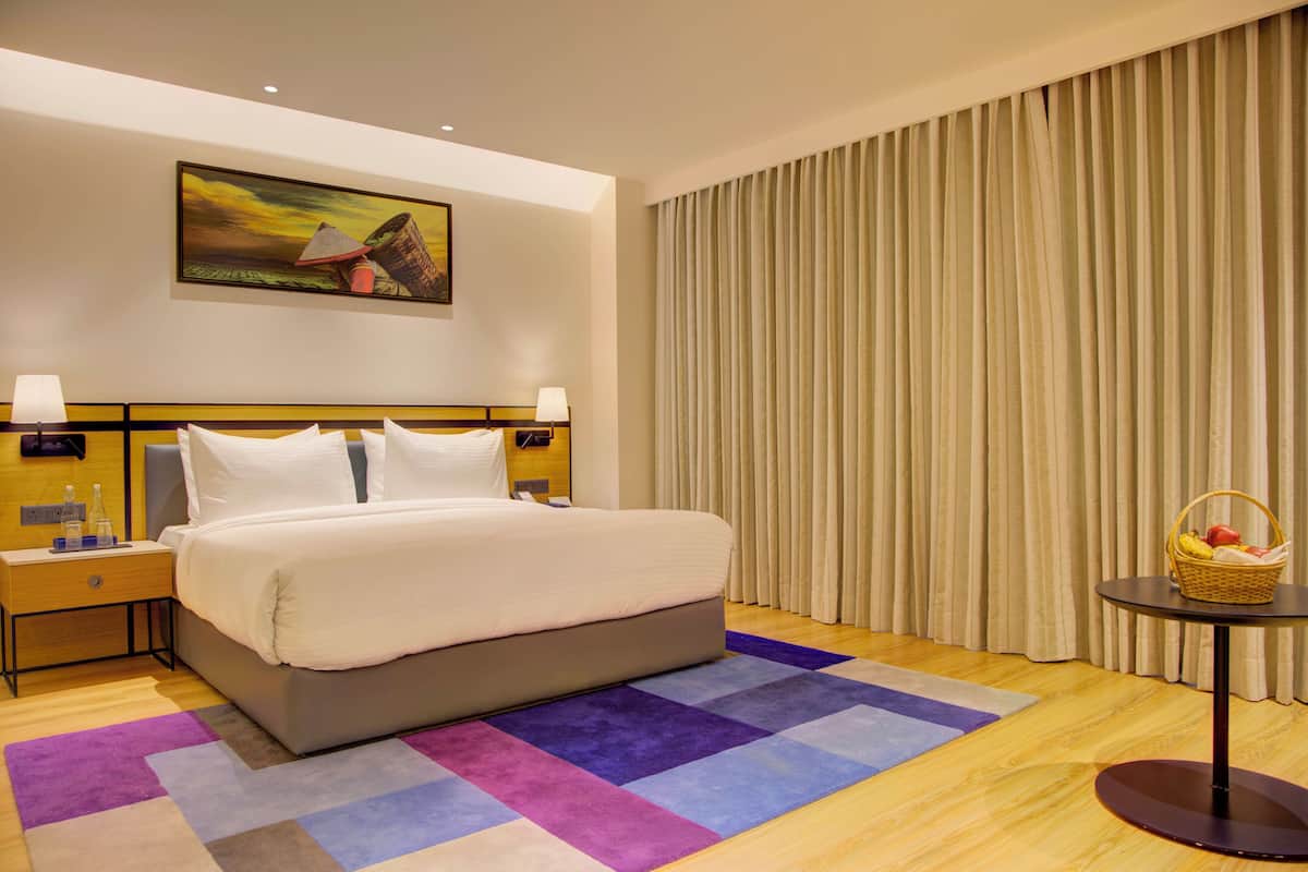 A hotel room with a large bed, two bedside tables with lamps, a colorful rug, a painting on the wall, and a window with curtains.