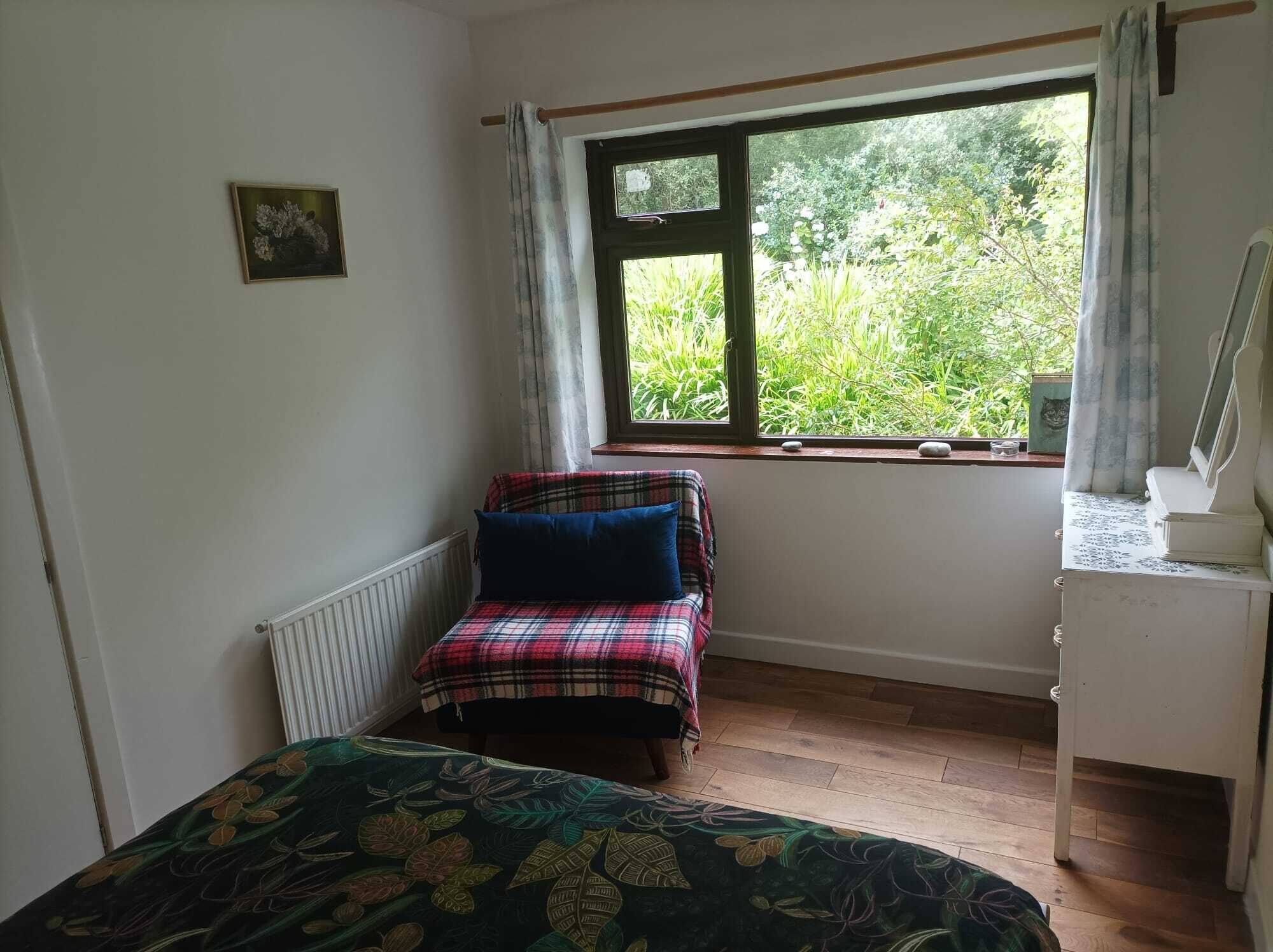 Une petite pièce agréable et chaleureuse, avec un lit simple, une fenêtre donnant sur un espace verdoyant, un radiateur et une commode avec un miroir.
