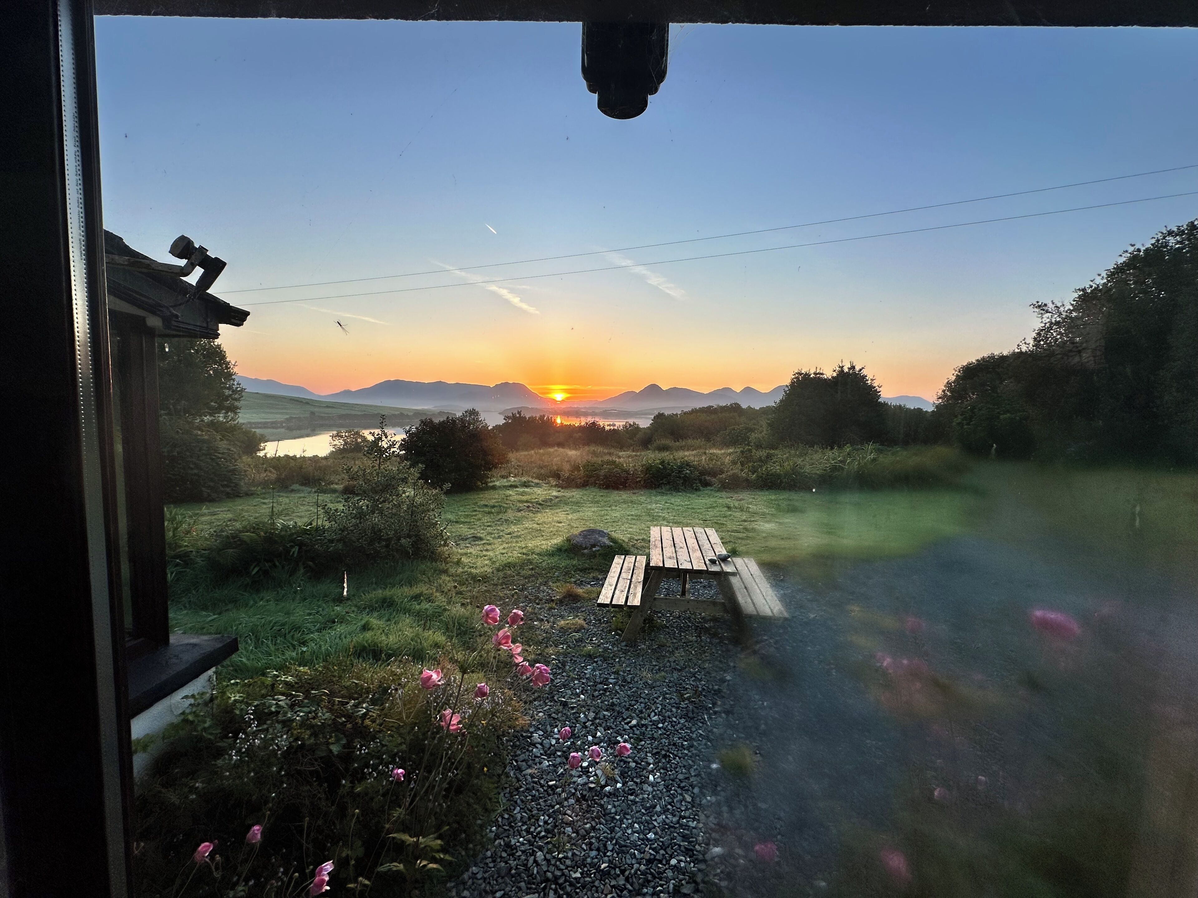 A view from a window overlooking a sunset, a grassy area with a wooden bench, and a body of water in the distance.