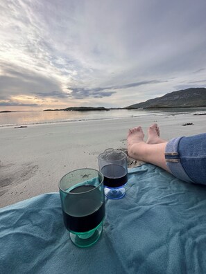 A beach scene with a blanket and two glasses of dark liquid, likely wine, on a sandy shore.