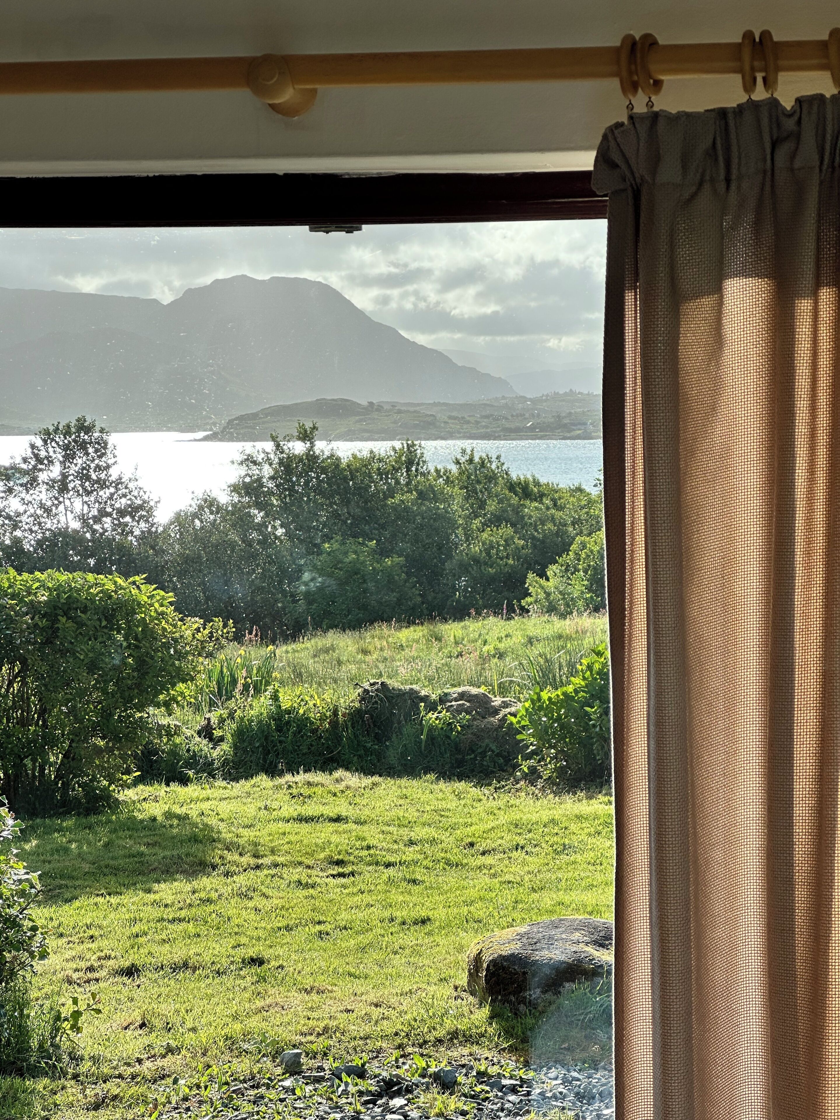 A room with a view of a lake and mountains through a window with a curtain.