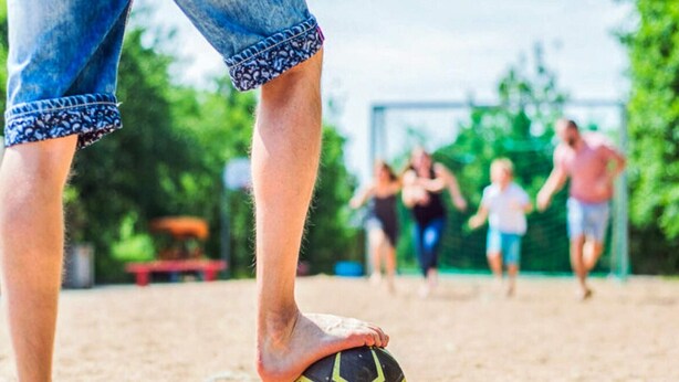 Iemand trapt een voetbal op een zandige ondergrond, met een doel in de verte.