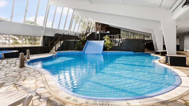 A swimming pool with a slide, stone flooring, and a view of the outdoors through large windows. - 6 Person Holiday Park Home in Hesselager (Hesselager)
