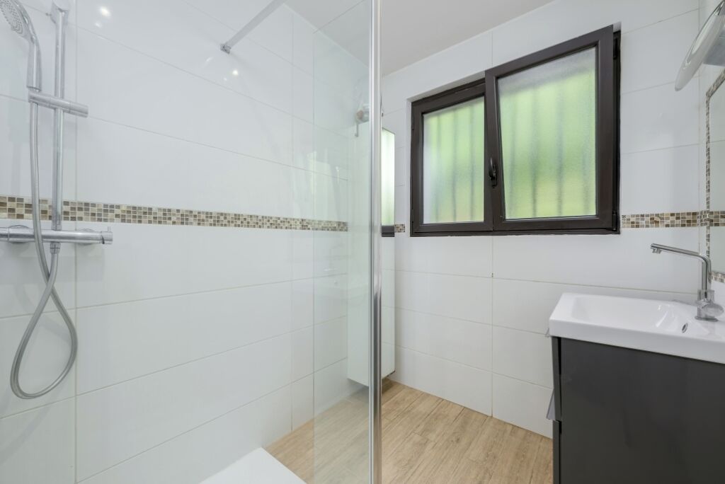A modern bathroom with white tiles, a glass shower enclosure, a wooden floor, and a window with blinds.