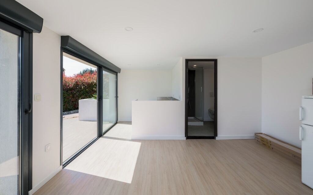 A modern, minimalist room with a large sliding glass door leading to an outdoor area, a small kitchen area, and a built-in bench.