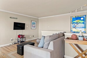 A living room with a TV, a sofa, a coffee table, a small table with a radio, and a wall art piece.