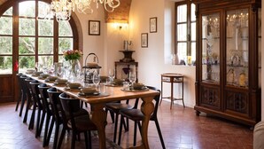 Comedor con una larga mesa dispuesta para una comida, rodeada de sillas. Un candelabro cuelga del techo, y un aparador con puertas de cristal exhibe diversos objetos.